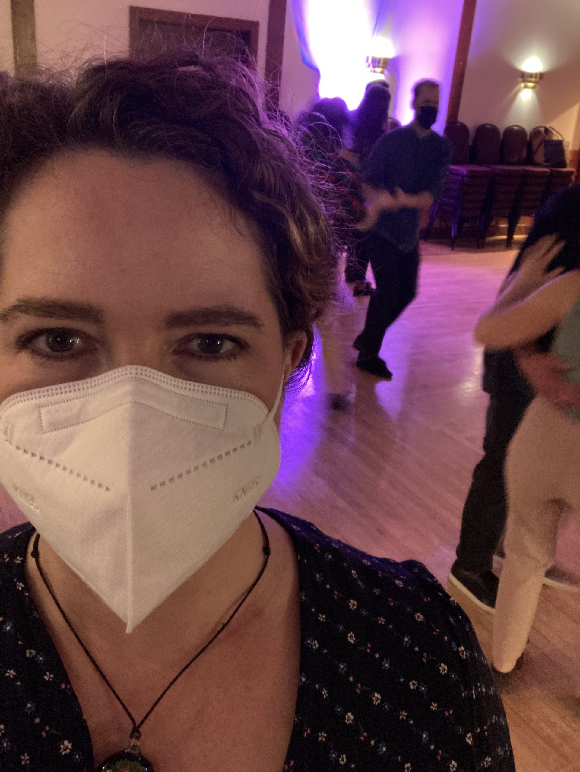 A selfie of Eve Rickert at a swing dance event. She looks into the camera wearing a white N95 mask and smiling. Masked people dance behind her.